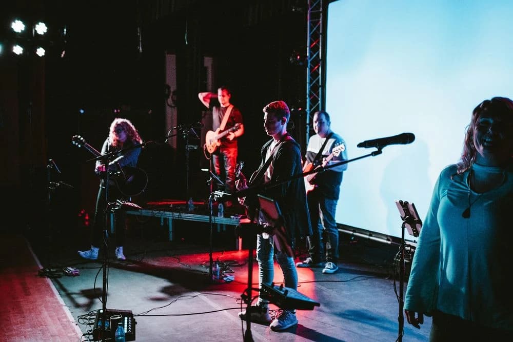 A band of five musicians playing guitars and bass on a stage with a large blue screen in the background.